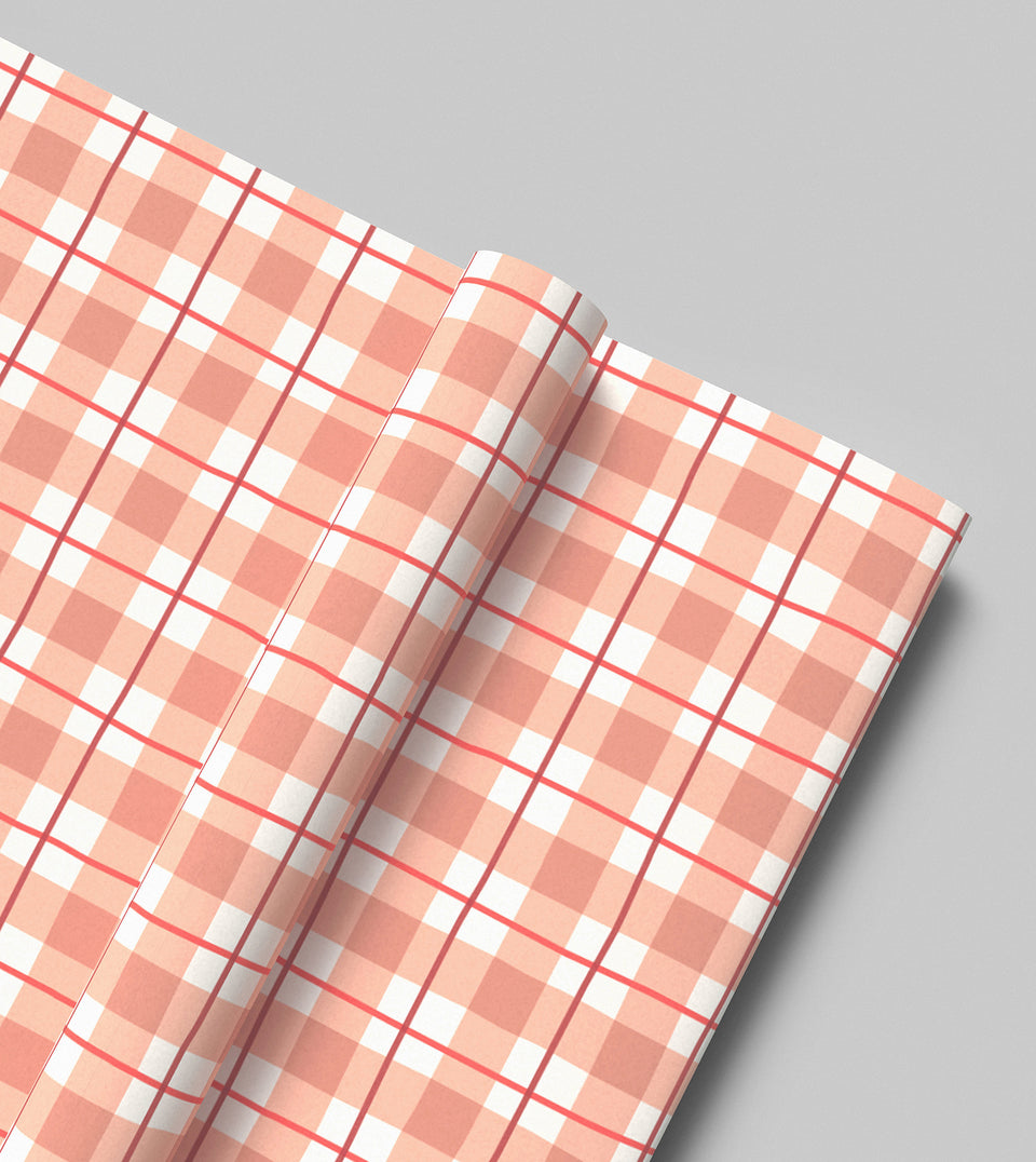 A roll of wrapping paper with a dusty rose, peachy pink, white, red and wine tartan plaid pattern, shown on a neutral gray background.