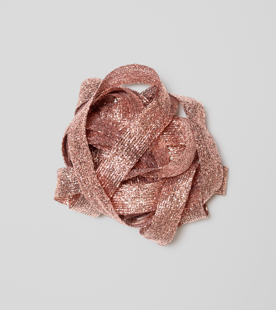 A coiled bundle of shimmering rose gold metallic braided ribbon on a solid light grey background. Studio Carta