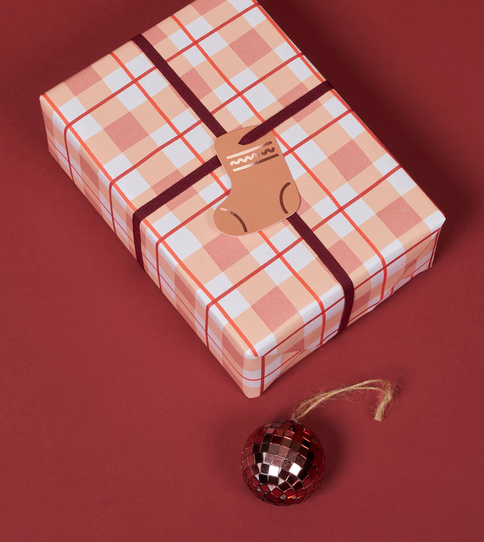 A wrapped rectangular gift box with a peachy pink, dusty rose, red, wine and white, plaid pattern, tied with a wine red ribbon and a metallic stocking gift tag, shown on a dark red background with a small mirrored ornament below.