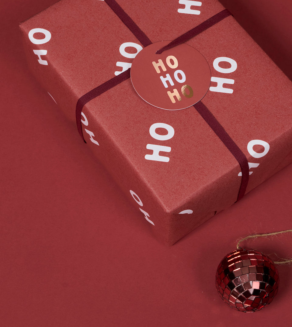 A square gift box wrapped in wine red festive paper printed with white 'HO HO HO' lettering, tied with a wine ribbon and topped with a round rift tag that reads 'HO HO HO' in metallic foil. A small red mirrored ornament hangs in the lower right corner against a matching red background.