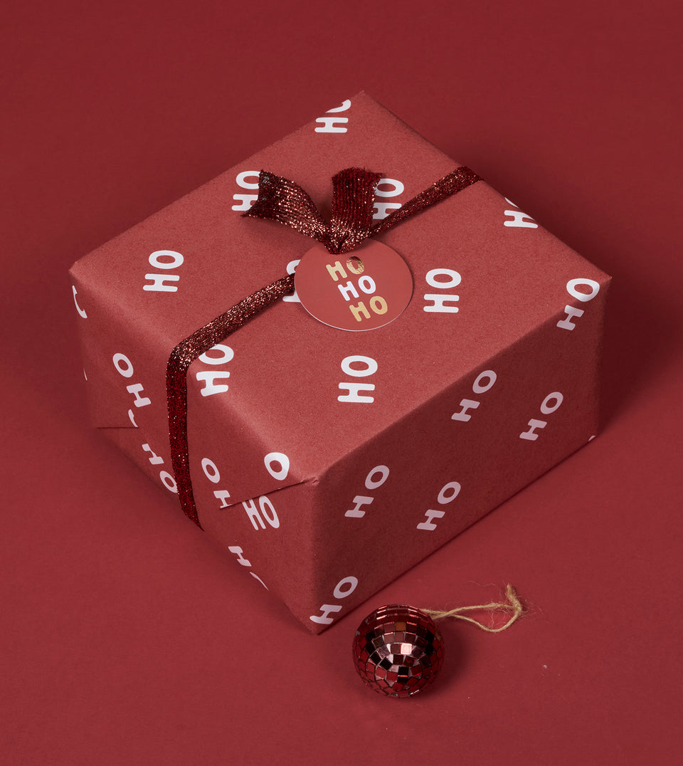 A square gift box wrapped in dark red paper with a white 'HO HO HO' pattern, tied with a glittery copper ribbon and bow, and topped with a round 'HO HO HO' gift tag on a matching red background with a small dark red ornament in front.