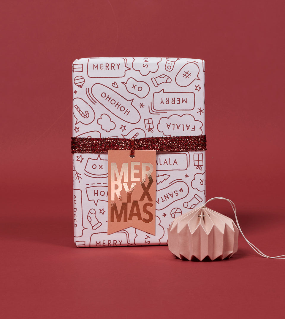 A wrapped gift box with festive Christmas-themed wrapping paper, metallic copper ribbon and a metallic 'Merry Christmas' tag, set against a solid dark red background with a small pink paper ornament nearby.