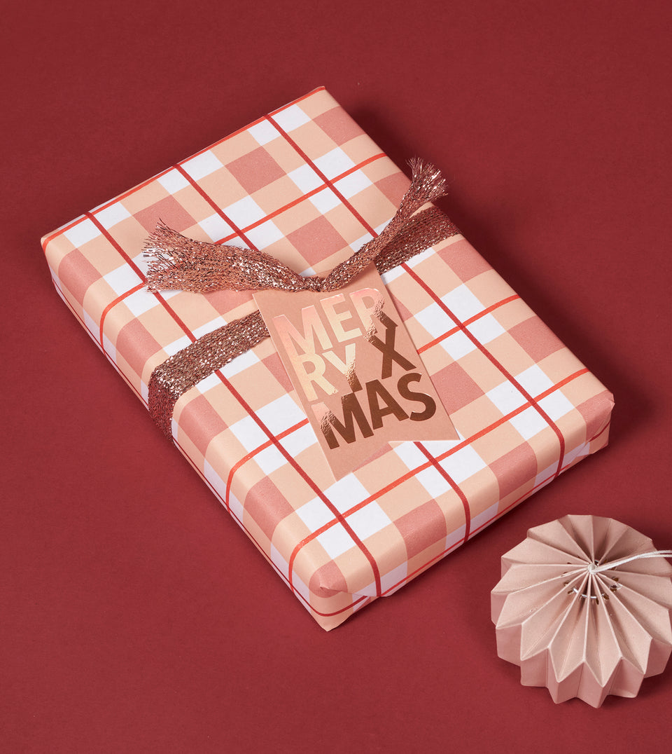 A wrapped rectangular gift box with peach, white, and red gingham wrapping paper, tied with a glittery metallic rose gold ribbon and a metallic dusty rose gift tag that reads “MERRY XMAS,” shown on a dark red background with a small pale pink ornament nearby.