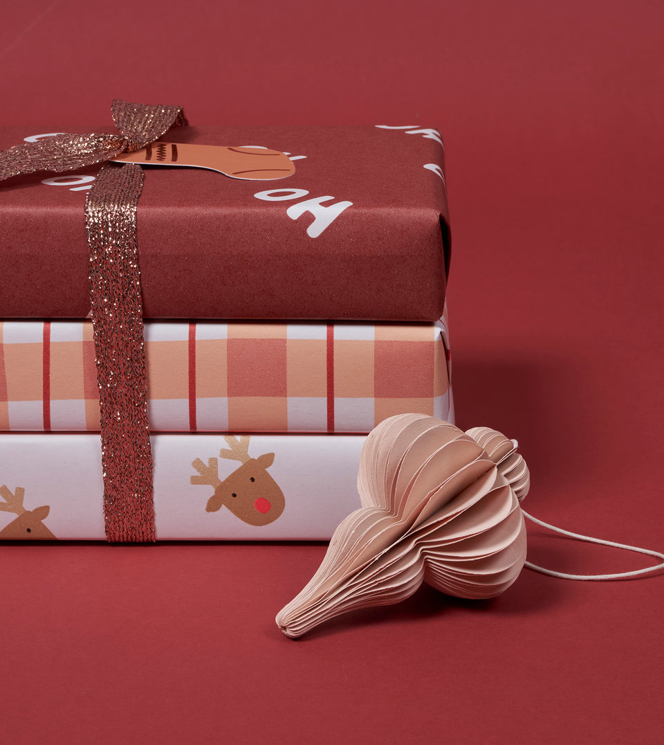 Three stacked Christmas-themed gift boxes wrapped in patterned paper, tied with a glittery rose-gold ribbon, with a beige paper ornament on a solid dark red background.