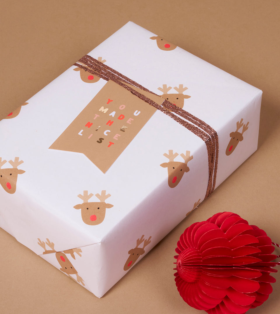 A square gift box wrapped in white paper printed with a repeating brown reindeer pattern, tied with a rose gold ribbon and a tan  tag that reads 'YOU MADE THE NICE LIST'. The box is photographed on a warm tan background with a red paper decoration in the lower right corner.