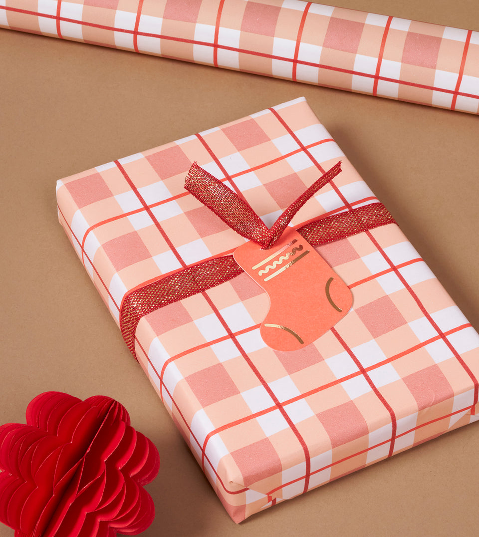 A wrapped gift box and a roll of matching plaid wrapping paper on a tan background, tied with a red ribbon and decorated with a red paper flower prop.