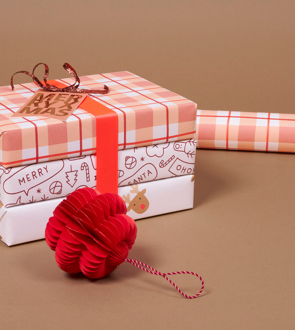 A stack of wrapped Christmas gift boxes with dusty rose, red and peach plaid wrapping paper, a red ribbon, and a metallic gift tag, alongside a red paper honeycomb ornament on a warm beige background.