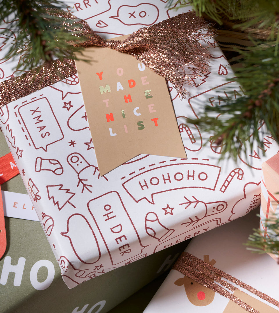 A wrapped Christmas gift box with white wrapping paper printed with wine red holiday line drawings, a glittery rose-gold ribbon, and a tan gift tag that reads 'YOU MADE THE NICE LIST' in multi-coloured letters. Pine branches frame the top of the scene.