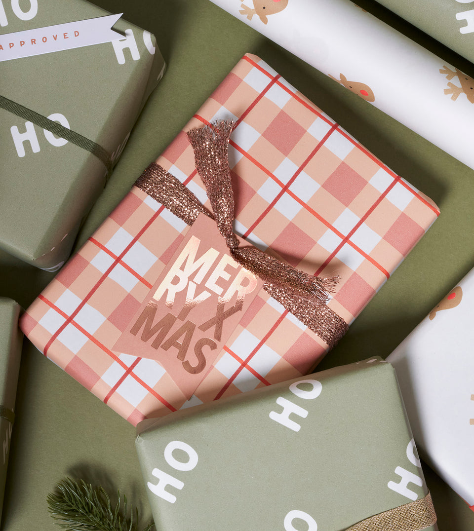 A wrapped gift box in the center with a peachy pink and red gingham pattern, tied with a glittering rose-gold ribbon and a hanging tag that reads 'MERRY XMAS'. Other wrapped presents in sage green and white wrapping paper are arranged around it on a green surface.