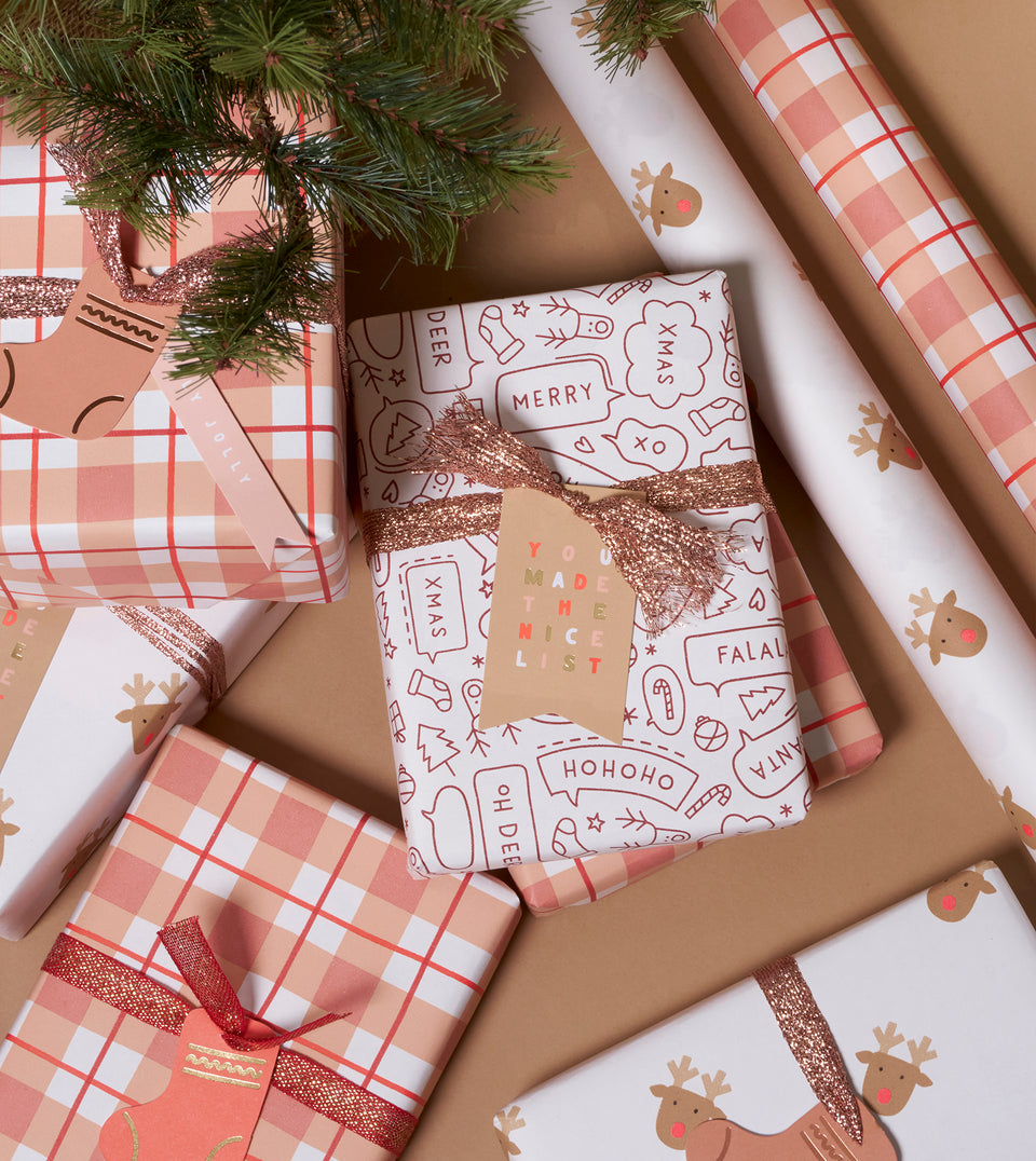 A styled flat-lay of Christmas gift wrapping paper and wrapped presents on a tan surface. The wrapping includes a plaid pattern in peachy pink, and red, and white paper with wine coloured holiday illustrations, and one roll with a reindeer print.