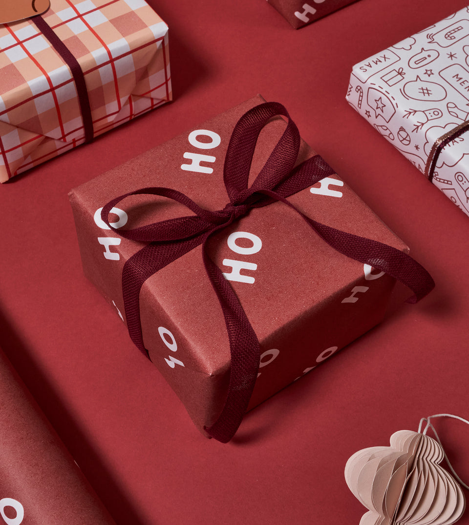 A wrapped gift box in wine red wrapping paper with white 'HO HO HO' text and a dark burgundy ribbon bow, styled on a matching red surface with other gift wraps partially visible.