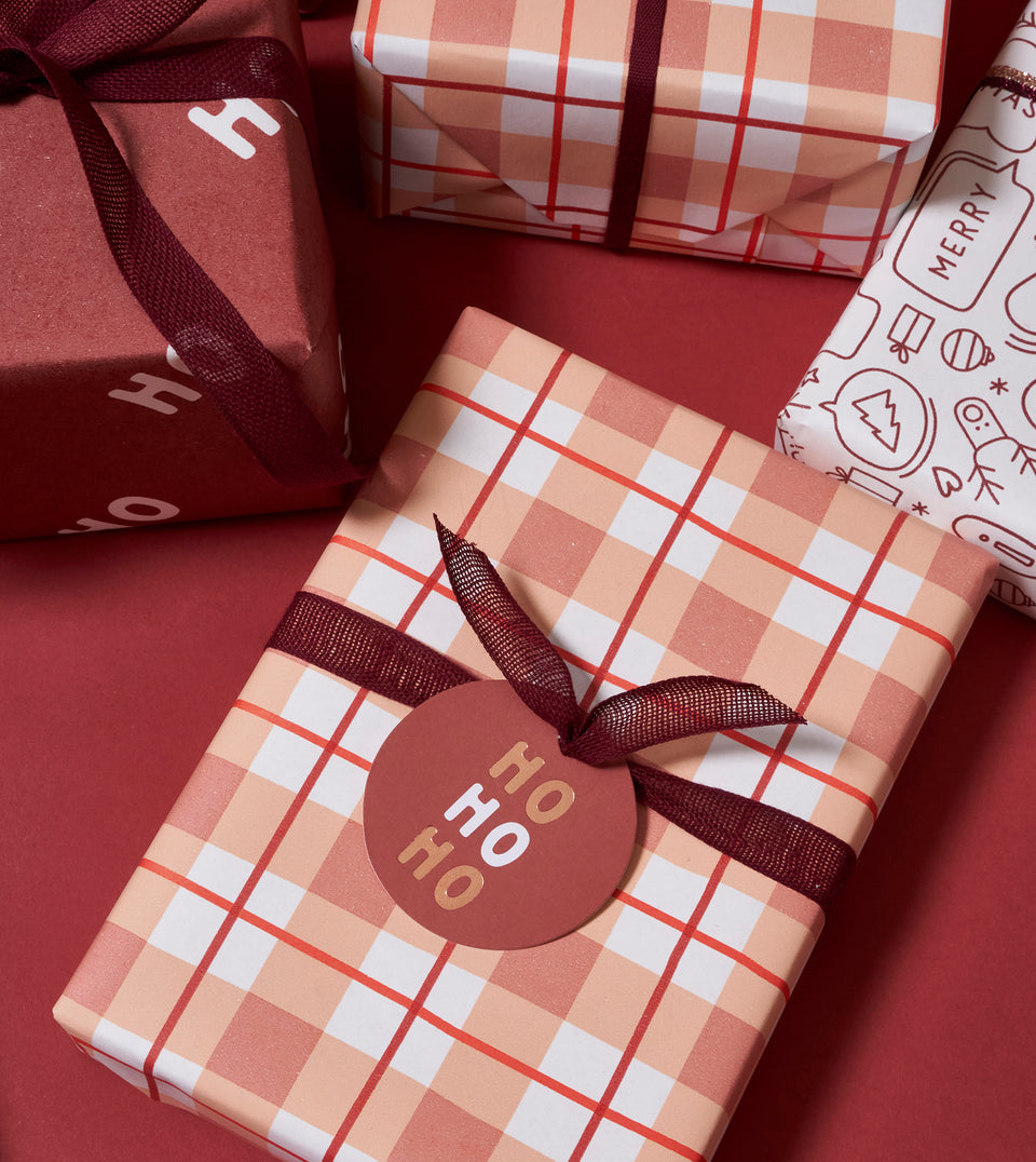 A wrapped gift box in the foreground with a peachy pink, white and red gingham pattern, tied with a dark burgundy ribbon and a round gift tag that reads 'HO HO HO'. Other holiday gift boxes are partially visible in the background on a solid red surface.