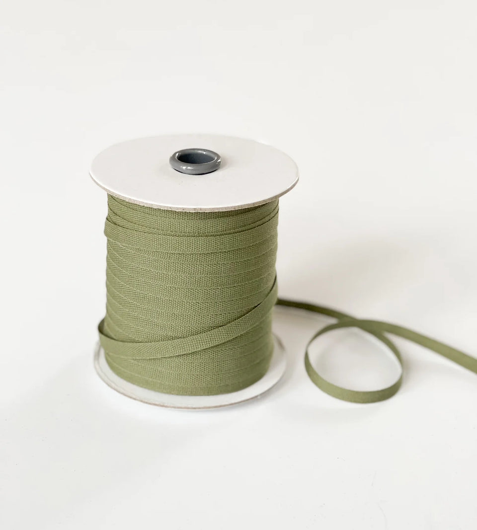 A spool of olive green ribbon with a white cardboard core, shown on a clean white background with a loose end of the ribbon trailing to the right. Courtesy of Studio Carta